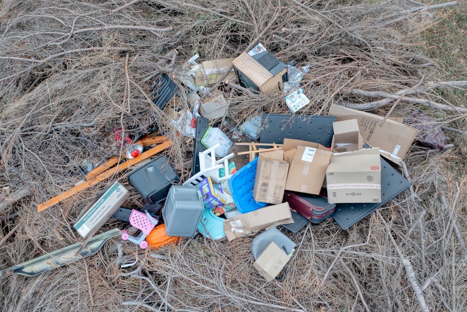 A pile of mixed household waste and clutter scattered across a dry, leafless outdoor area. The debris includes various cardboard boxes, some flattened and others partially crushed, with visible logos