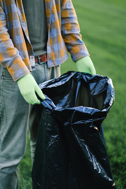 A person standing outdoors on a grassy area, wearing a plaid shirt and gray trousers, is holding open a large black rubbish bag lined with silver material, prepared for waste collection. The individua
