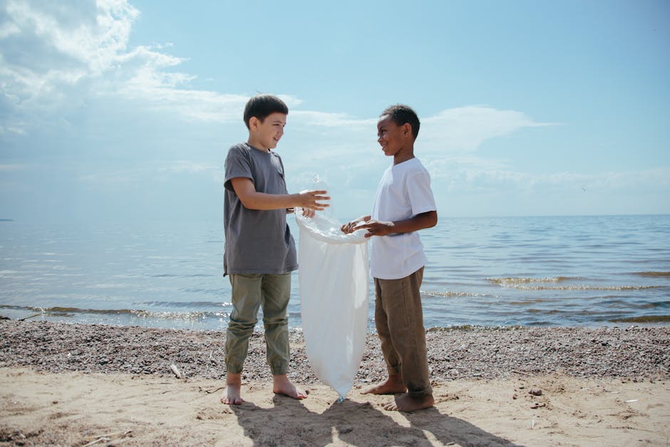 Two young boys with short dark hair and medium skin tone are standing barefoot on a sandy beach near the water's edge. One boy, wearing a grey T-shirt and khaki pants rolled up to his calves, is holdi