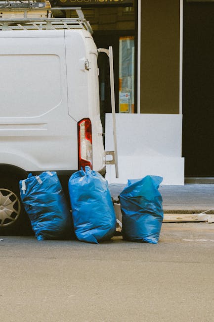 A waste collection operative operates a large red refuse collection vehicle on a street, with the rear hopper open to load waste materials. The worker, dressed in a high-visibility vest with orange an